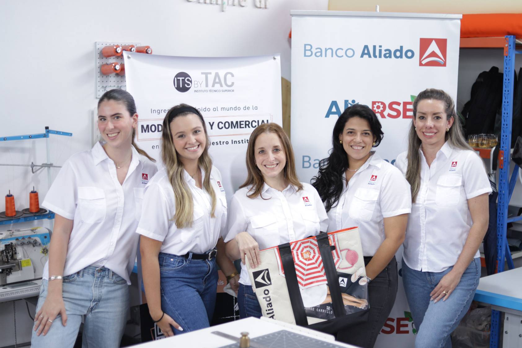 Un grupo de colaboradoras con camisas corporativas posa sonriente sosteniendo un bolso de material reciclado frente a pancartas de Banco Aliado, Al rse e ITS by TAC en un taller textil.